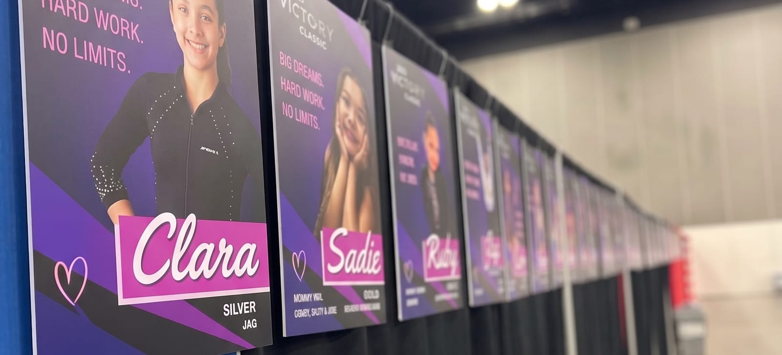 A long row of FanBoards clipped to the drape along a competition floor, each with an athlete's photo and first name.